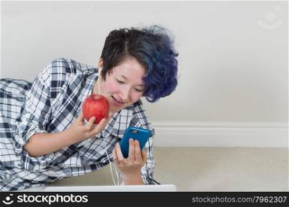 Teen girl holding apple while looking at cell phone with computer in forefront while lying down listening to music at home.