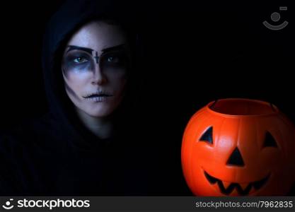 Teen girl in scary face paint holding large pumpkin container on black background. Trick or treat concept for Halloween with selective lighting.