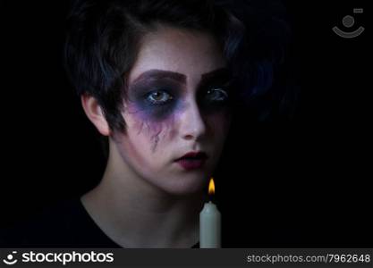 Teen girl in scary makeup holding candle with flame on black background.