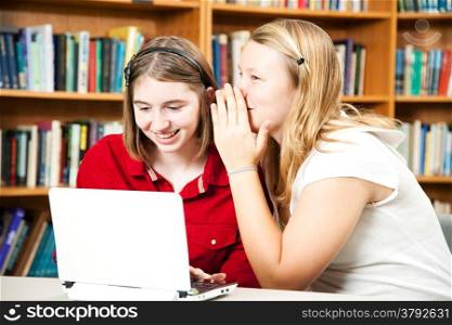 Teen girl students in the library whispering secrets and using the computer.