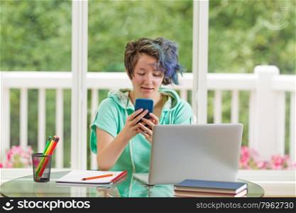 Teen girl texting while doing her homework. Large windows in background with blurred out bright green trees and flowers.