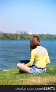 teen girl with a laptop sitting on a meadow near the river