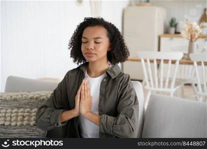 Teenage african american girl is practicing yoga at home. Curly young spanish woman is sitting on couch in lotus pose with her eyes closed. Harmony and balance. Wellness and spirituality.. Teenage african american girl is practicing yoga at home. Woman is sitting on couch in lotus pose.