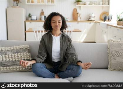 Teenage afro girl is practicing yoga at cosy home. Young woman sitting on couch in lotus pose. Concentration and balance. Wellness and emotional wellbeing, tranquility and recreation, stress relief.. Teenage afro girl is practicing yoga at cosy home. Wellness and emotional wellbeing, stress relief.