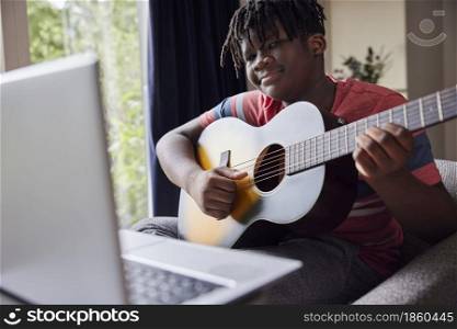 Teenage Boy Learning To Play Acoustic Guitar With Online Lesson On Laptop Computer At Home