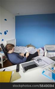 Teenage Boy Sitting At Desk In Bedroom Using Digital Tablet