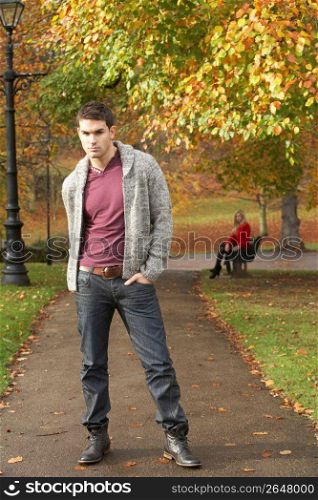 Teenage Boy Standing In Autumn Park With Female Figure On Bench In Background