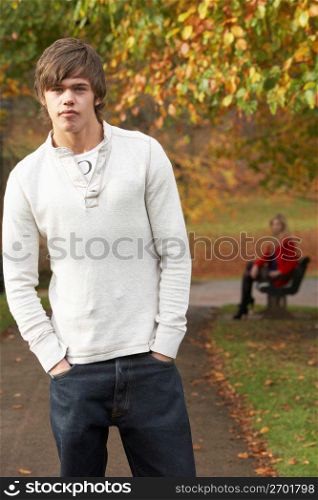 Teenage Boy Standing In Autumn Park With Female Figure On Bench In Background