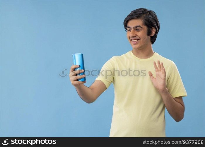 Teenage boy waving hand during video call on Smartphone against blue background