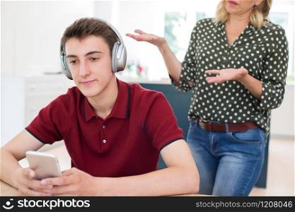 Teenage Boy Wearing Headphones And Using Mobile Phone Being Nagged By Mother At Home