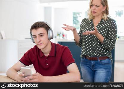 Teenage Boy Wearing Headphones And Using Mobile Phone Being Nagged By Mother At Home