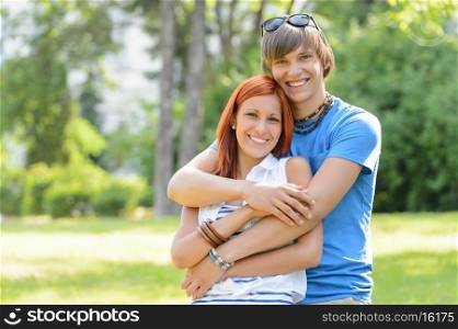 Teenage couple hugging in sunny park smiling at camera