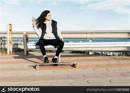 Teenage female sitting on a bench with her feet on the longboard in front of the sea