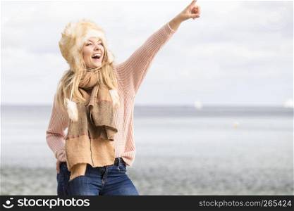 Teenage female spending time outdoor on the beach during autumn weather pointing at copy space. Wearing sweater, scarf and fur hat.. Fashionable woman on beach pointing