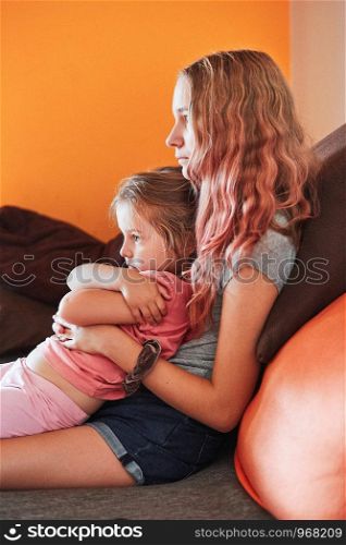 Teenage girl and her little sister watching TV together, sitting concentrated on sofa at home. Real people, authentic situations