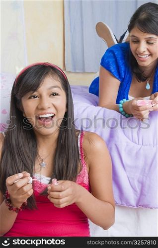Teenage girl applying nail polish on her nails with a young woman lying on the bed behind her