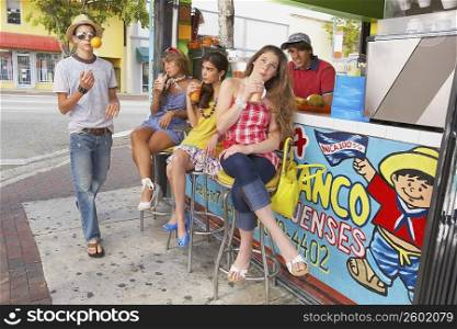Teenage girl drinking juice with her two friends and a teenage boy tossing a lemon in front of them