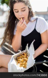 Teenage Girl Eating French Fries Sitting On Bench Outdoors