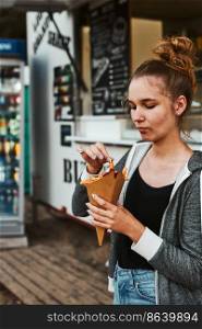 Teenage girl eating potato fries from carton cone standing in front of food truck. Teenager having a fast food meal outdoors during summer vacations. Teenage girl eating potato fries standing in front of food truck during summer vacations