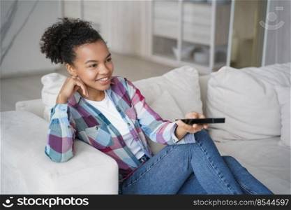 Teenage girl enjoying tv series watching and shifts channels with remote controller. Afro young woman has leisure in morning and sitting on couch on quarantine. Reality show watching.. Teenage girl enjoying tv series watching and shifts channels. Afro young woman has morning leisure.