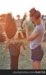 Teenage girl helping to water the flowers growing in flower pot, pouring water from green watering can, working in backyard at sunset. Candid people, real moments, authentic situations