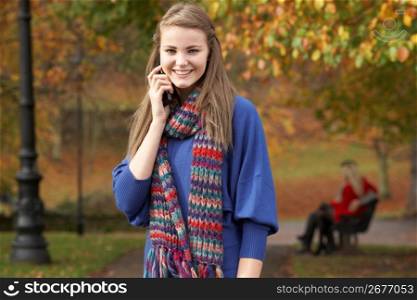 Teenage Girl On Mobile Phone In Autumn Park With Couple On Bench In Background