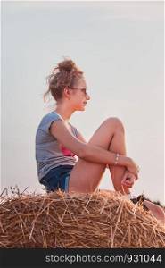 Teenage girl sitting on a hay bale at sunset relaxing while spending summertime in the village. Candid people, real moments, authentic situations