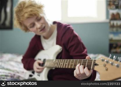 Teenage Girl Sitting On Bed Learning To Play Electric Guitar In Bedroom