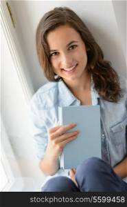 Teenage girl student sitting by window holding book smiling at camera
