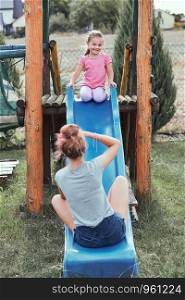 Teenage girl taking a photo her younger sister in a home playground in a backyard. Happy smiling sisters having fun on a slide together on summer day. Real people, authentic situations