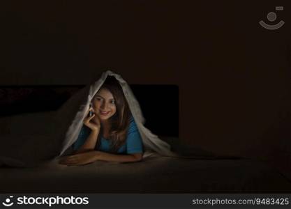 Teenage girl talking on her mobile phone under a blanket. 