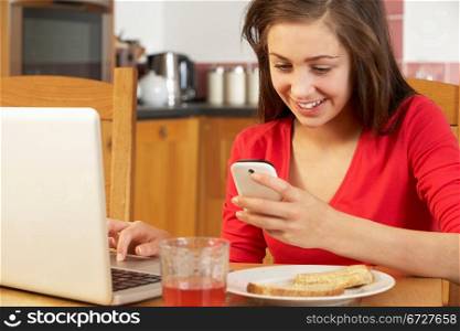 Teenage Girl Using Laptop And Mobile Phone Whilst Eating Breakfast