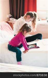 Teenage girl using mobile phone together with her little sister watching animated movie on tablet, both girls sitting in bed in bedroom