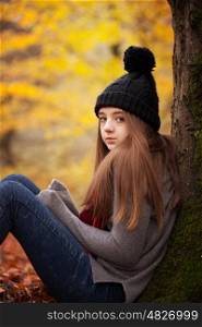 Teenage girl wearing a woolen hat sitting beside a tree with autumnal colours in the background