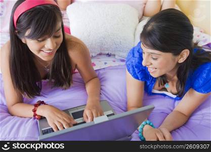 Teenage girl working on a laptop with a young woman lying beside her on the bed