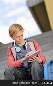 Teenage student boy using tablet sitting on steps outside school