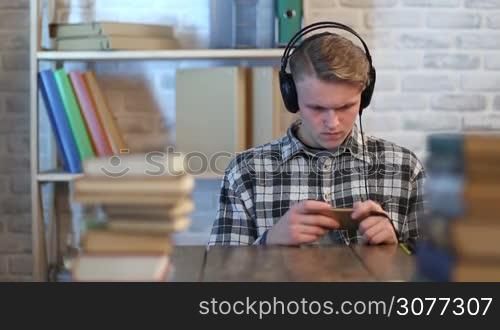 Teenage student with headphones taking online learning course in his smartphone at home. Attractive young man browsing cellphone and listening to music with headphones while having a break from studying.