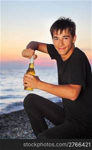 teenager boy opens beer bottler on stone seacoast in evening