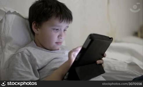 Teenager boy using touch pad lying in bed leaning on the pillow. Entertainment with tablet PC