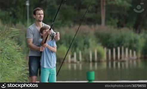 Teenager learning how to fish with spinning rod and reel with the help of his father at the pond. Dad and his son casting fishing rod together and enjoying leisure on the lake in countryside. Slow motion.