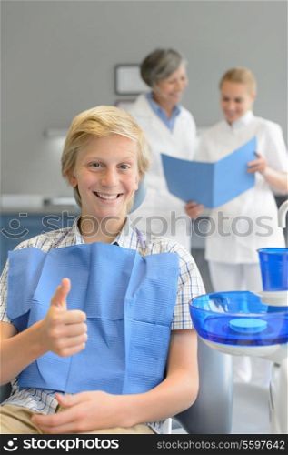 Teenager patient sitting at dental surgery thumbup dentist assistant background