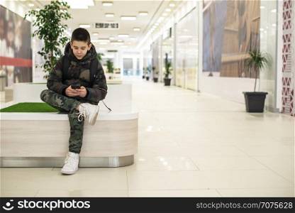 Teenager playing with smartphone in modern commercial center. Bench and flower in a pot. Modern building interior. Technology and communication concept with child in contemporary building interior.