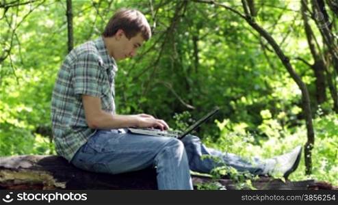 teenager sits on log with laptop.