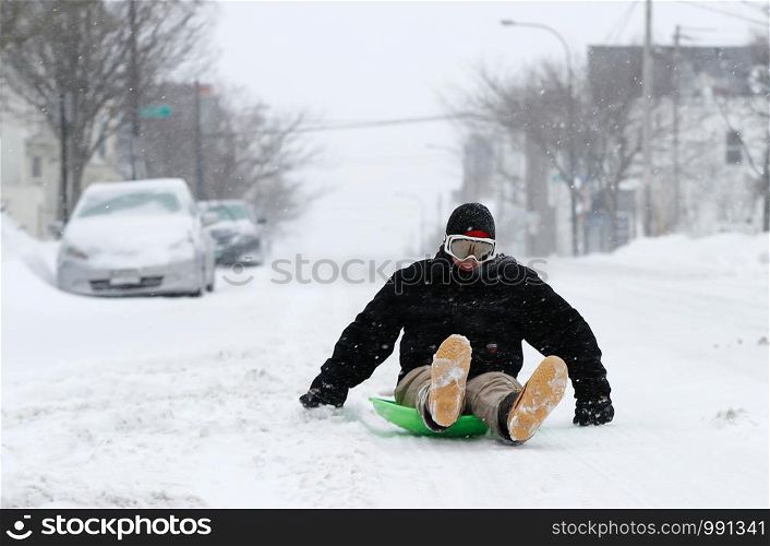 Teenager slides on a snowy winter road on a sled. Along the roadside much snow cars swept the snow and gray house. Gabby DiGiacomo