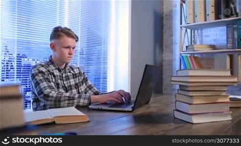 Teenager studying in library using laptop. Happy student with the winning gesture working on computer. Attractive hipster sitting at the desk working on pc gesturing excited on victory and success, expression of triumph.