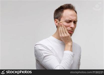 Teeth problem or dental illness. Close up of male feeling pain, holding his cheek with hand and suffering from bad toothache, isolated on gray background, free space. Sensitive enamel concept