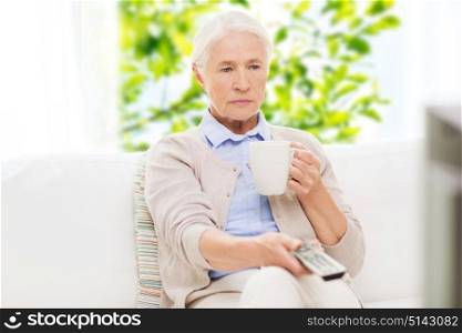television, age and people concept - senior woman watching tv, drinking tea and changing channels by remote control at home over green natural background. senior woman watching tv and drinking tea at home