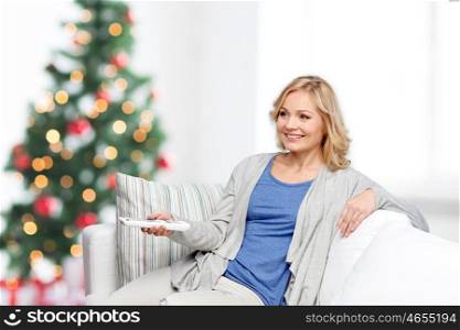 television, holidays, leisure and people concept - smiling woman sitting on couch with tv remote control at home over christmas tree lights background