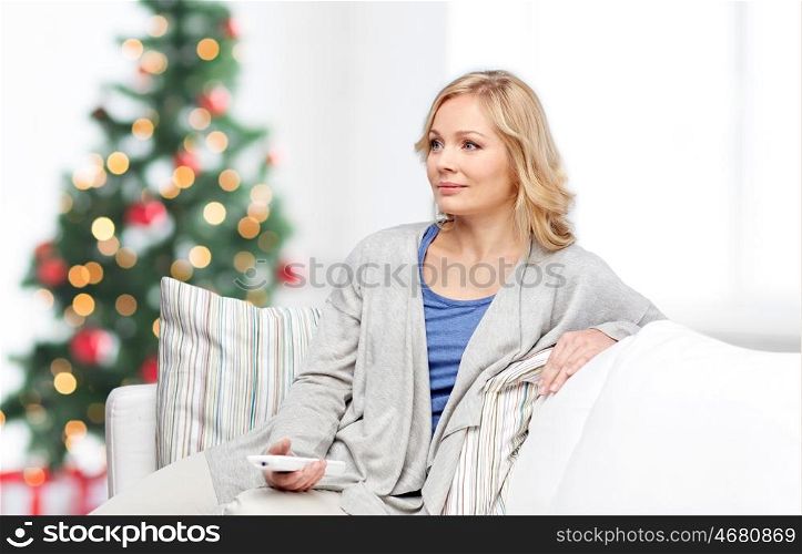 television, holidays, leisure and people concept - smiling woman sitting on couch with tv remote control at home over christmas tree lights background