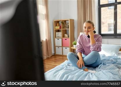 television, mass media and people concept - happy smiling girl with remote control watching tv at home. happy smiling girl watching tv at home